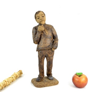 May include: A hand-crafted ceramic figurine of a person with a thoughtful expression, standing on a brown base. The figurine is painted in shades of brown and has a realistic appearance.