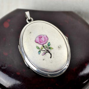 May include: Oval silver locket pendant with a pink rose design. The locket features a hinged design and a small loop for a chain. The rose has green leaves and a black stem. The locket is set against a dark background.