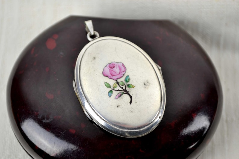 May include: Oval silver locket pendant with a pink rose design. The locket features a hinged design and a small loop for a chain. The rose has green leaves and a black stem. The locket is set against a dark background.