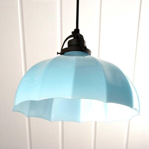 May include: A light blue pendant lamp with a scalloped shade. The lamp hangs from a black cord and fixture. The shade is a soft, pastel blue and has a rounded, umbrella-like shape. The background is white.