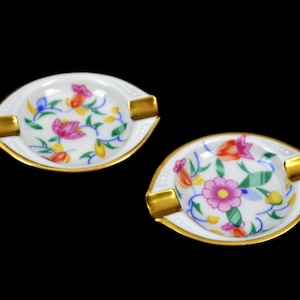 Pode incluir: Duas cinzeiros de porcelana branca com acabamentos dourados e desenhos florais. Cada cinzeiro apresenta um padrão colorido de flores rosa, laranja e azul com vinhas verdes. Os cinzeiros têm um pequeno descanso retangular dourado.