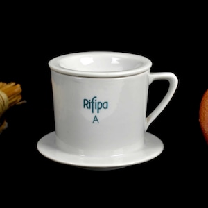 May include: A white ceramic Vietnamese coffee filter with a handle and a saucer base. The side of the filter features the blue text Rifipa A. To the left is a bundle of light brown dried herbs, and to the right is a red and yellow apple.