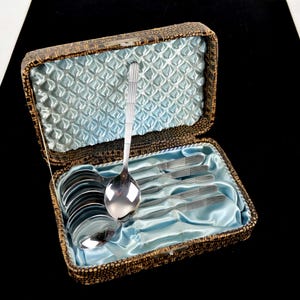 Könnte beinhalten: Ein Vintage-Besteckset in einer dekorativen Schatulle. Das Set enthält Löffel und Gabeln, alle aus glänzendem Silbermetall. Die Schatulle ist braun mit einem blauen Satinfutter, und das Besteck ist ordentlich darin angeordnet.