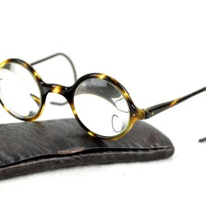 May include: Vintage round eyeglasses with a tortoise shell pattern on the frames, resting on a dark brown leather case. The lenses are clear, and the temples are thin and dark.