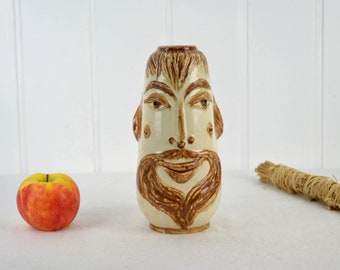 May include: A ceramic vase shaped like a face with a brown beard and mustache. The vase is cream colored with brown details.