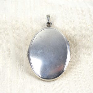 May include: A silver-colored oval locket with a small loop at the top for a chain. The locket has a shiny, reflective surface and is set against a cream-colored fabric background. The locket is closed.