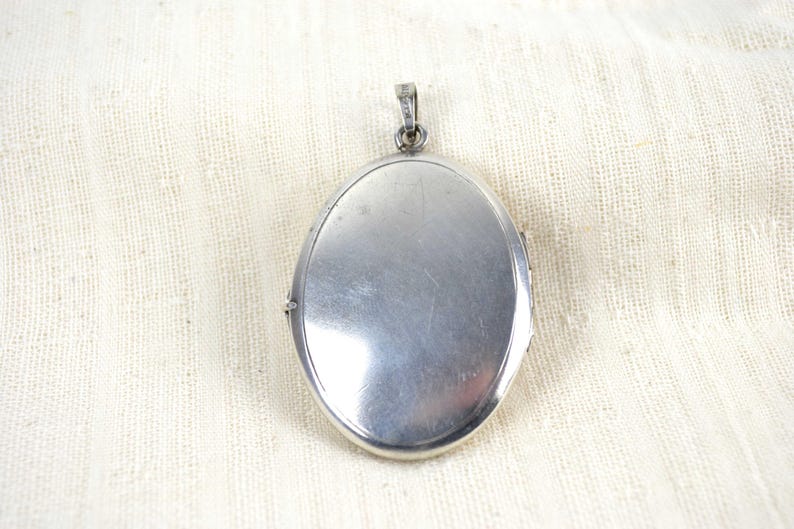 May include: A silver-colored oval locket with a small loop at the top for a chain. The locket has a shiny, reflective surface and is set against a cream-colored fabric background. The locket is closed.