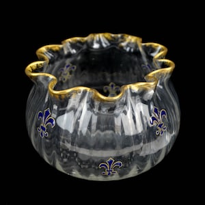 Puede incluir: Un cuenco de vidrio transparente con un borde dorado y diseños de flores de lis azules. El cuenco tiene un borde festoneado y es perfecto para servir aperitivos o postres.