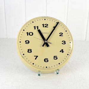 Puede incluir: Un reloj automático Kienzle vintage de color crema con números y manecillas negros. El reloj tiene una esfera redonda y se muestra sobre un soporte transparente. La esfera del reloj tiene una cubierta de vidrio.