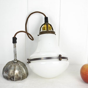May include: A white glass pendant light fixture with a metal frame and a brown cord. The light fixture is hanging from a white ceiling. There is a red apple on the table below the light fixture.
