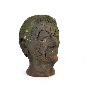 May include: A ceramic sculpture of a human head with a cracked and weathered surface, covered in moss and dirt. The head is tilted slightly to the side, and the eyes are closed.