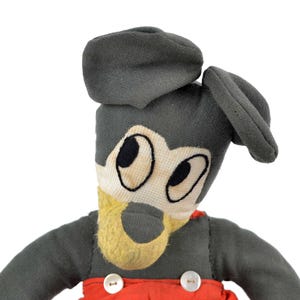 May include: A vintage plush toy of Mickey Mouse wearing a grey cap and red overalls. The toy has black eyes and a yellow nose.