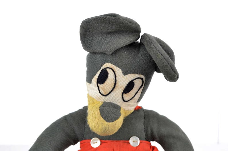 May include: A vintage plush toy of Mickey Mouse wearing a grey cap and red overalls. The toy has black eyes and a yellow nose.