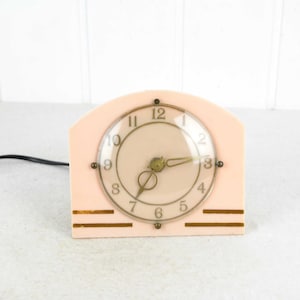 Könnte beinhalten: Eine vintage rosafarbene elektrische Uhr mit goldenen Akzenten und einem klaren Zifferblatt. Die Uhr hat römische Ziffern und ein schwarzes Kabel.