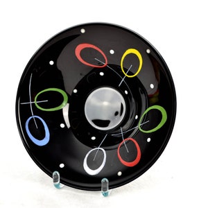 May include: Black glass bowl with a white, red, yellow, green, and blue abstract design. The bowl has a slight curve to the rim.