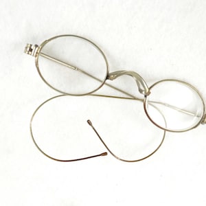 Könnte beinhalten: Antike silberfarbene Brille mit runden Gläsern. Die Brille hat einen filigranen Drahtrahmen und ein einzigartiges Brückendesign. Die Bügel sind gebogen. Die Gläser sind klar.