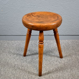 May include: A wooden stool with three turned legs and a round seat. The stool is made of light brown wood and has a rustic appearance.
