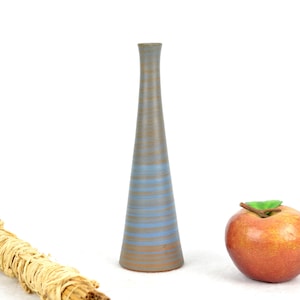 May include: A blue and brown ceramic vase with a striped pattern. The vase is tall and narrow, with a rounded base. It is sitting on a white surface, next to a bundle of dried straw and a red apple.