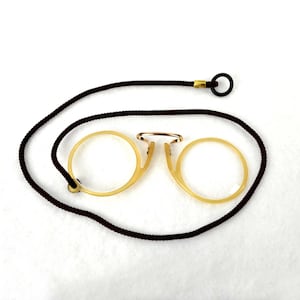 Puede incluir: Gafas antiguas de pinza con un cordón marrón oscuro. Las gafas tienen una montura de color amarillo claro y un puente dorado. El cordón tiene un lazo en el extremo. Las gafas están sobre un fondo blanco.