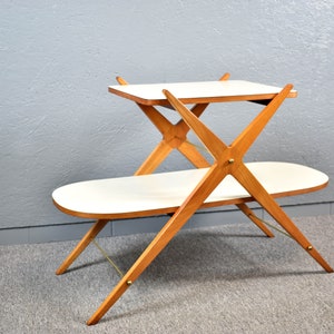 May include: A two-tiered wooden serving cart with a white top and a white oval-shaped bottom shelf. The cart has a unique X-shaped design with brass accents.