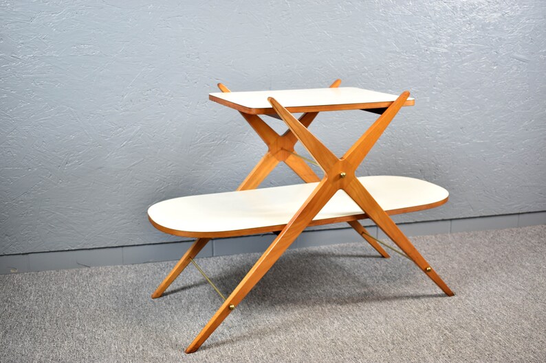 May include: A two-tiered wooden serving cart with a white top and a white oval-shaped bottom shelf. The cart has a unique X-shaped design with brass accents.