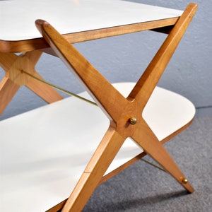May include: A two-tiered wooden serving cart with a white laminate top and bottom. The cart has a unique X-shaped base with brass accents.