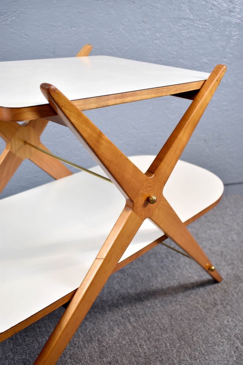May include: A two-tiered wooden serving cart with a white laminate top and bottom. The cart has a unique X-shaped base with brass accents.