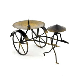 May include: A metal candle holder shaped like a person pulling a cart. The cart has a round, gold-colored top and two wheels. The person is wearing a conical hat.