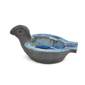 Puede incluir: Un cenicero de cerámica con forma de pájaro. El cenicero es principalmente gris oscuro con un interior y borde azules. La cabeza del pájaro está erguida y el cenicero tiene un acabado liso y brillante.