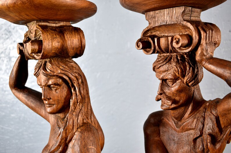 Puede incluir: Dos esculturas de madera talladas intrincadamente de figuras sosteniendo una plataforma circular. Las esculturas est&aacute;n hechas de madera marr&oacute;n oscura y presentan expresiones faciales detalladas y cabello elaborado.