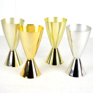 Könnte beinhalten: Vier goldene und silberne Metall-Schnapsgläser mit einem ausgestellten Boden. Die Gläser sind in einer Reihe auf einer weißen Oberfläche angeordnet.