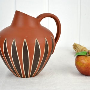 May include: A terracotta pitcher with a brown and white geometric design. The pitcher has a wide base and a narrow neck with a handle.