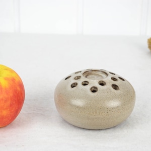 May include: A small, round, light gray ceramic vase with a ring of holes around the top. The vase is sitting on a white surface.