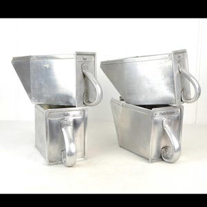 Peut inclure: Quatre récipients vintage en aluminium avec des poignées. Chaque récipient a une base rectangulaire et un dessus trapézoïdal. Les récipients sont étiquetés avec du texte, dont "Salz" et "Hale-Rocker".