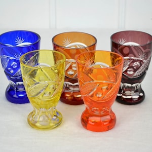 May include: Five small, stemmed, cut-glass shot glasses in different colors: blue, yellow, orange, red, and purple.