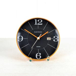 May include: A black and orange analog clock with a date display. The clock face has black hands and white numbers. The clock face also has the word "Electronic" printed on it.