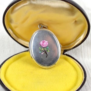 May include: Oval silver locket with a pink rose design, set in a brown case with a yellow velvet interior. The locket is open, revealing the interior of the case. The locket is a vintage piece.