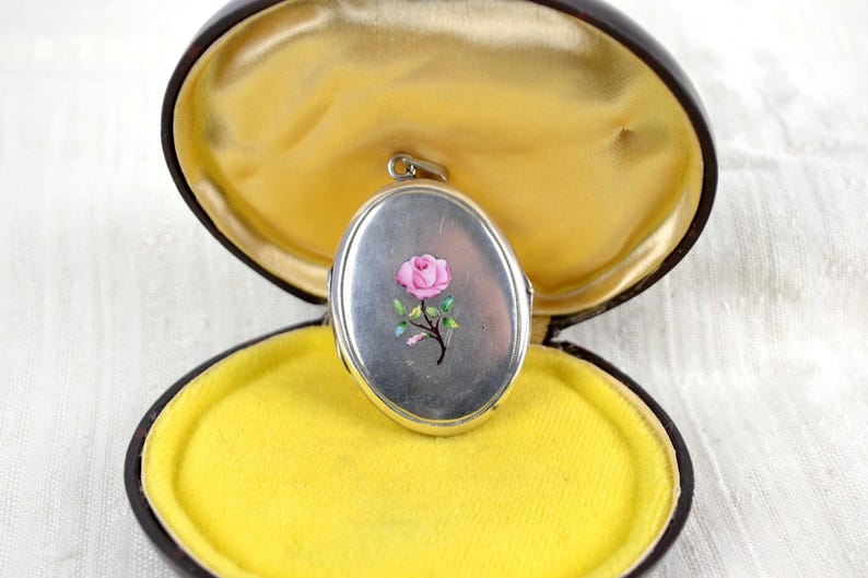 May include: Oval silver locket with a pink rose design, set in a brown case with a yellow velvet interior. The locket is open, revealing the interior of the case. The locket is a vintage piece.