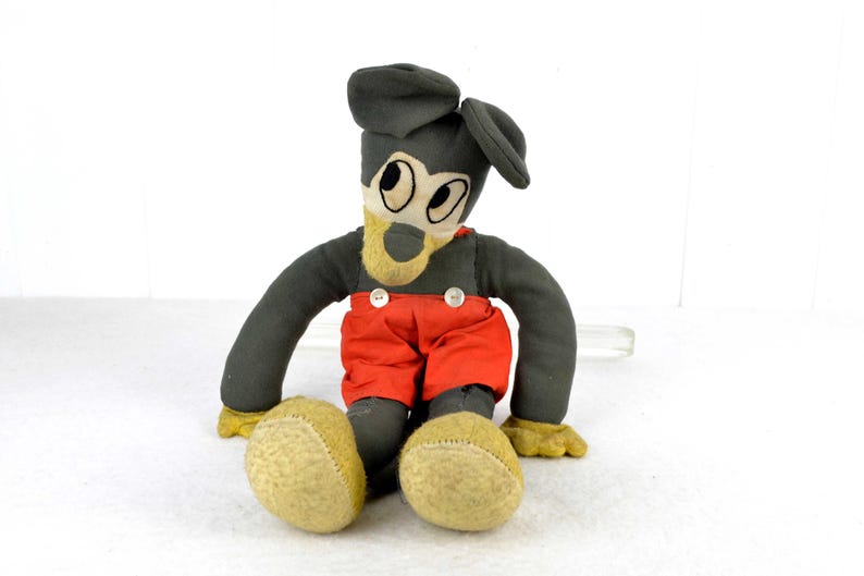 May include: A vintage plush toy of Mickey Mouse, with a grey body, red trousers, and black shoes. The toy has a black felt hat and large, white eyes.
