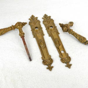 Puede incluir: Conjunto de herrajes de puerta de latón antiguo, que incluye dos placas de puerta ornamentadas y dos manijas de puerta a juego. Las manijas tienen detalles intrincados y un diseño decorativo.