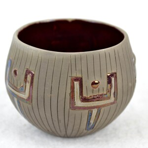 May include: A small, round ceramic bowl with a dark red interior and a light grey exterior. The bowl has a geometric design in shades of red, gold, and blue.
