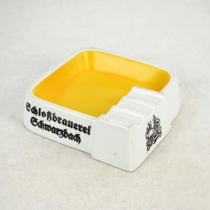 Op de afbeelding: Witte asbak van keramiek met een gele binnenkant en een zwarte inscriptie die "Schloßbrauerei Schwarzbach" luidt.