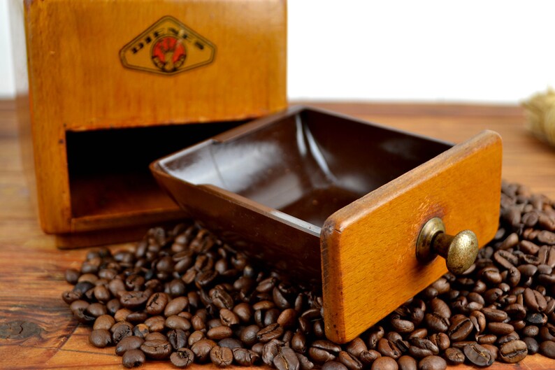 K&ouml;nnte beinhalten: Eine Kaffeem&uuml;hle aus Holz mit einer Schublade, die ge&ouml;ffnet ist und mit Kaffeebohnen gef&uuml;llt ist. Die Schublade hat einen Messingknopf. Die M&uuml;hle hat ein Etikett mit der Aufschrift "DREH-MEH".