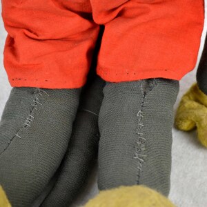 May include: Close-up of a vintage Mickey Mouse plush toy. The toy is wearing red shorts and has black felt legs with visible stitching. The toy's hands are yellow felt.