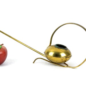 May include: A gold-colored metal watering can with a long spout and a circular handle. The watering can is on a white surface.