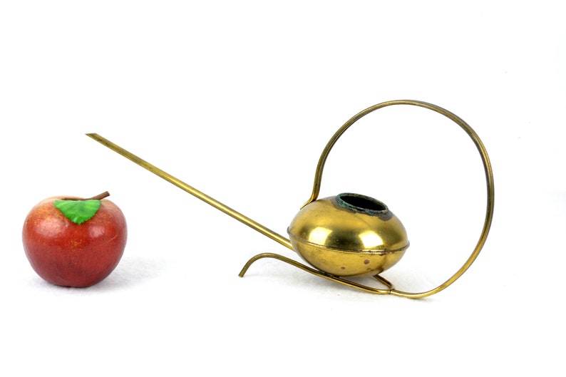 May include: A gold-colored metal watering can with a long spout and a circular handle. The watering can is on a white surface.