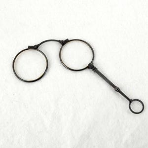 Könnte beinhalten: Antike Pince-Nez-Brille aus schwarzem Metall. Die Brille hat zwei runde Gläser, die durch einen Steg und einen Griff mit einem kleinen Ring verbunden sind. Das Design ist schlicht und elegant, passend für historische oder Vintage-Accessoires.