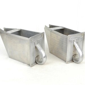 Peut inclure: Deux récipients en aluminium brossé avec des poignées incurvées. Les récipients ont une forme rectangulaire avec un design trapézoïdal. Les poignées sont fixées sur le côté des récipients. Les récipients sont vides et reposent sur une surface blanche.