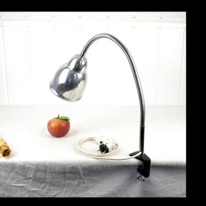 May include: A silver clamp-on desk lamp with a flexible gooseneck and a metal shade. The lamp is plugged in and has a white cord.