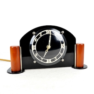 May include: An Art Deco style clock with a round face and black hands. The clock has a black base with two brown cylindrical accents on either side.